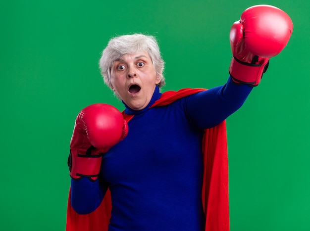 Supereroe donna anziana che indossa mantello rosso con guantoni da boxe guardando la telecamera spaventata e preoccupata in piedi su sfondo verde