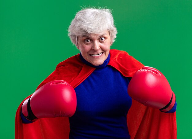 Senior woman superhero wearing red cape with boxing gloves looking at camera strained and excited standing over green