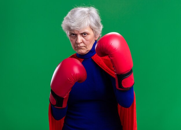 Free photo senior woman superhero wearing red cape with boxing gloves looking at camera with serious confident expression ready to fight standing over green background