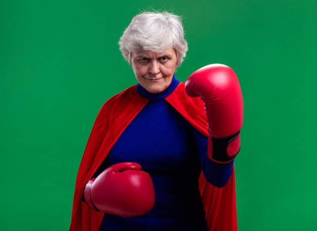 Старшая женщина-супергерой в красной накидке с боксерскими перчатками смотрит в камеру с серьезным уверенным выражением лица, готовая к бою