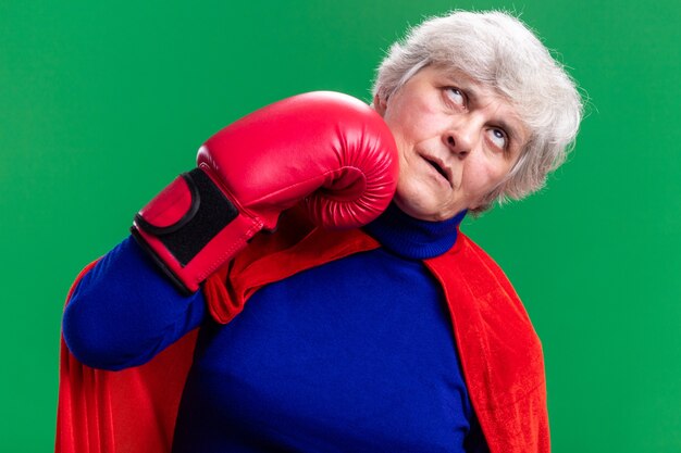 Free photo senior woman superhero wearing red cape with boxing gloves punching herself standing over green background