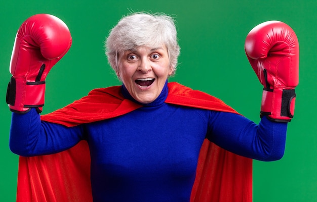 Senior woman superhero wearing red cape with boxing gloves raising hands happy and excited standing over green background