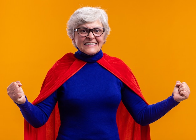 Senior woman superhero with glasses wearing red cape looking at camera happy and excited clenching fists standing over orange