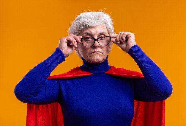 Senior woman superhero with glasses wearing red cape looking at camera with serious face standing over orange background