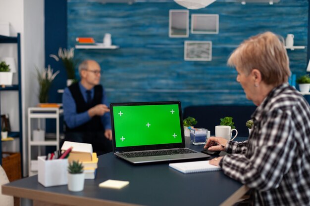 Senior woman taking notes on notebook looking at portable computer with copy space available. Elderly woman working on laptop with green screen and husband holding tv remote control.