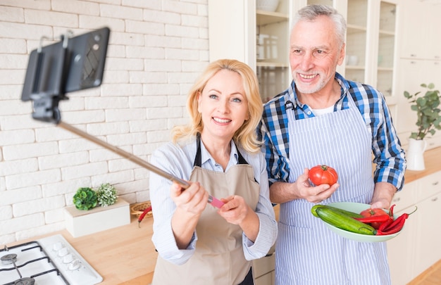 Старшая женщина принимая selfie на мобильном телефоне при его супруг держа овощ в руке
