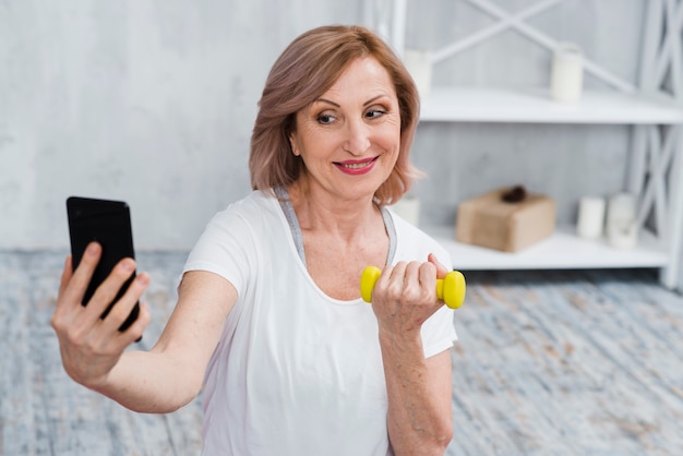 Старшая женщина принимая selfie с гантелями в руке