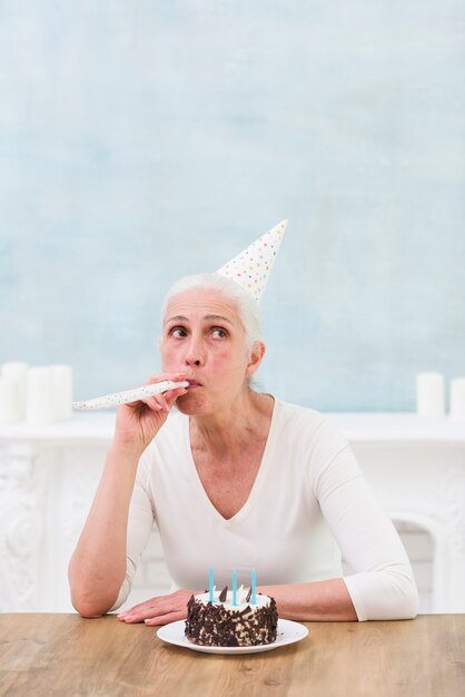 Senior woman wearing hat blowing party horn with tasty cake and candles on wooden table