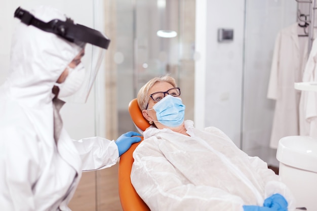 コロナウイルスの間に口腔病学のオフィスで化学防護服を着ている年配の女性。歯科医院での診察中に防護服を着た年配の女性。