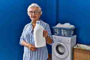 Free photo senior woman with grey hair holding detergent bottle looking positive and happy standing and smiling with a confident smile showing teeth