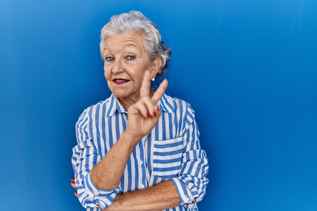 Пожилая женщина с седыми волосами, стоящая на синем фоне, улыбающаяся со счастливым лицом, подмигивающая в камеру, делая знак победы номер два
