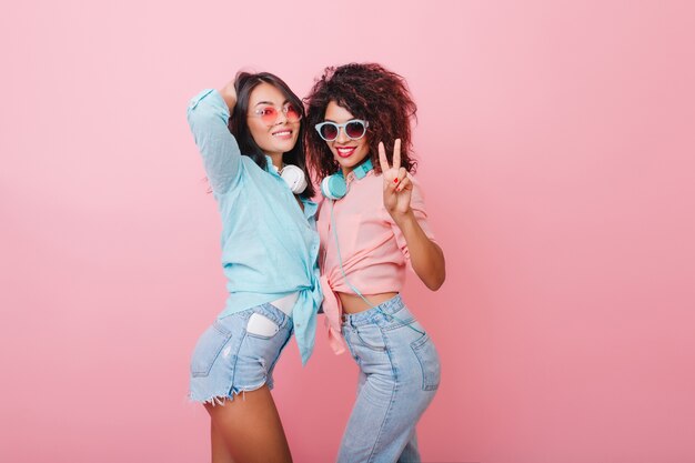 Sensual african girl with trendy curly hairstyle standing in carefree pose beside asian friend. Cute european lady in blue shirt touching her hair while posing with mulatto woman.