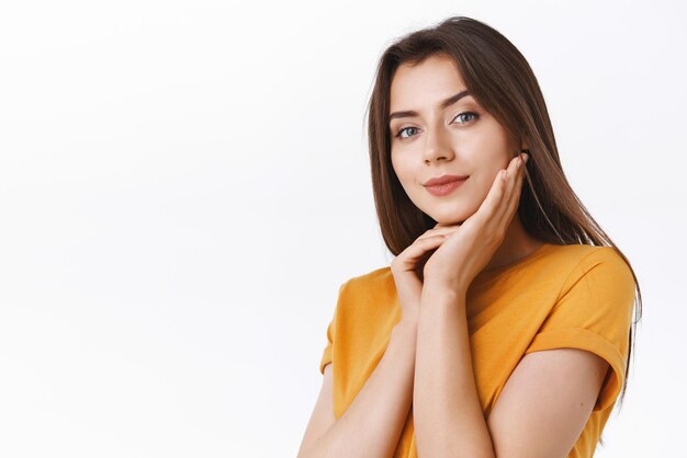 Sensual attractive sassy brunette woman in yellow tshirt gently touching clear skin smiling and gazing camera coquettish express sexy charming attitude standing alluring over white background