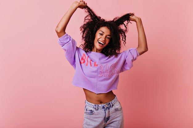 Sensual black girl posing on rosy with happy face expression. Jocund african woman having fun in studio with pink interior.