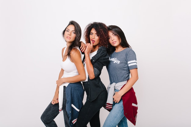 Sensual black woman in skinny jeans posing gently touching latin female friend. Indoor portrait of lovely asian girl in gray t-shirt standing next to university mates.