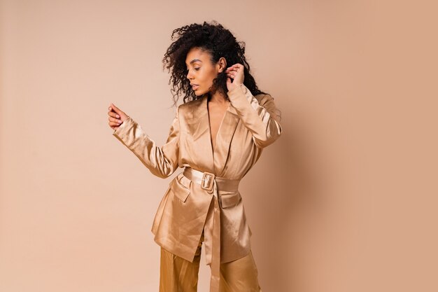 Sensual black woman with beautiful wavy hairs in elegant  golden satin suit posing over beige wall. Spring fashion look.