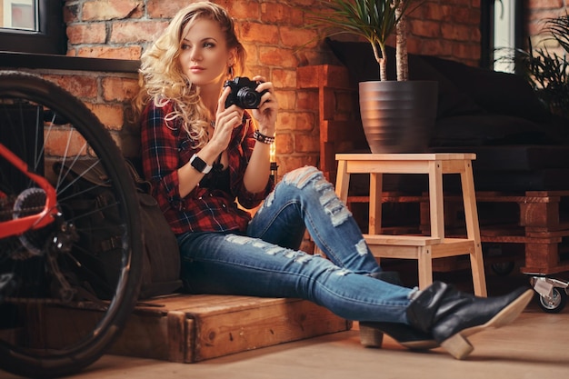 Sensual blonde hipster girl with long curly hair dressed in a fleece shirt and jeans holds a camera sitting on a wooden box at a studio with a loft interior.