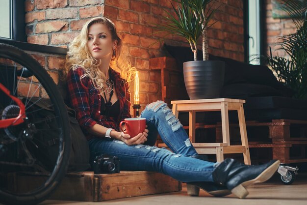 Sensual blonde hipster girl with long curly hair dressed in a fleece shirt and jeans holds a cup of morning coffee sitting on a wooden box at a studio with a loft interior, looking away.