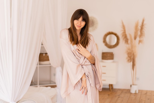보헤미안 스타일의 세련된 밝은 침실에서 베이지색 실크 기모노를 입고 포즈를 취하는 관능적인 브루네트 여성.