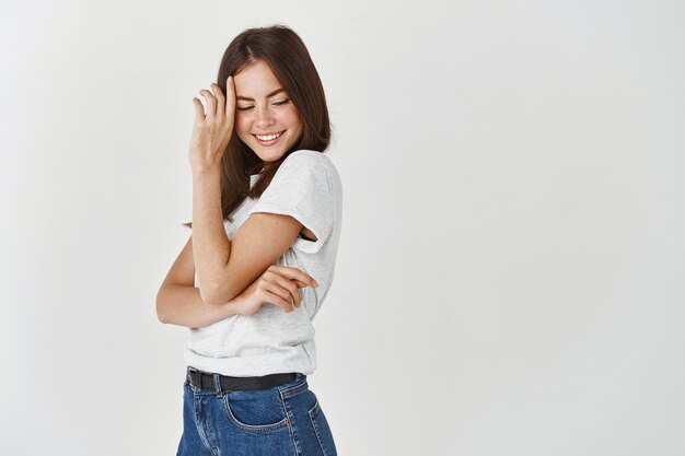 Sensual brunette woman standing in profile, blushing and smiling, looking down coquettish, standing over white wall