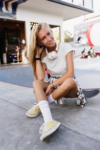 Sensual caucasian woman in white t-shirt sitting on longboard. Outdoor photo of carefree blonde lady playing with her hair in skate park.