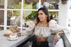 Free photo sensual confident woman in stylish clothes enjoying coffee coffeeshop attractive female model sitting cafe having lunch with admirer smiling look away pleased breakfast at outdoor restaurant