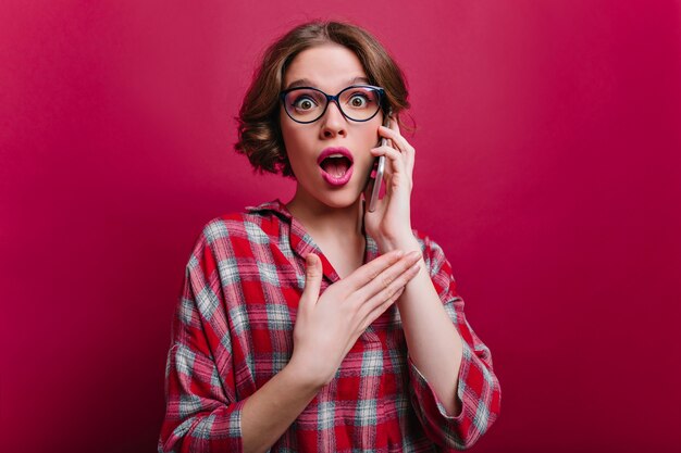 Sensual dark-eyed girl in trendy shirt talking on phone with surprised face expression. Indoor portrait of glamorous young woman holding smartphone and expressing amazement.