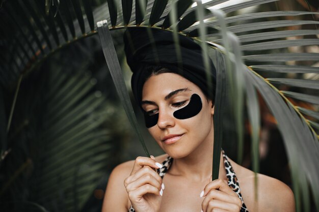 Sensual female model with eye patches posing near palm tree leaves. Outdoor shot of appealing woman in turban standing on nature background.