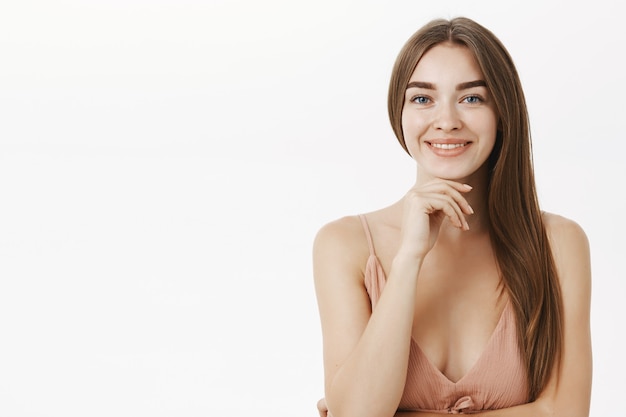 sensual and feminine brunette in cute beige dress holding hand on chin and smiling broadly having pure clear skin delighted to have no problems using minimal makeup over grey wall