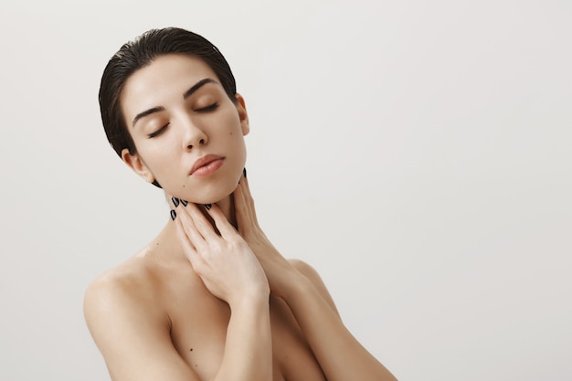 Sensual feminine woman standing naked with closed eyes, touching neck gently