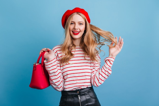 Sensual french girl playing with blonde hair.  smiling young lady in beret isolated on blue wall.