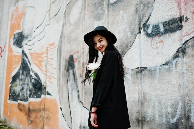 Sensual girl all in black red lips and hat Goth dramatic woman hold white chrysanthemum flower against graffiti wall