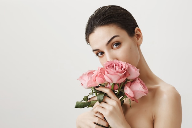 Sensual girlfriend smelling bouquet of roses while standing naked on grey