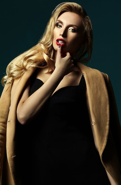 古典的な黒の衣装と唇に触れるオーバーコートで新鮮なメイクと美しい金髪の女性モデルの女性の官能的な魅力の肖像画
