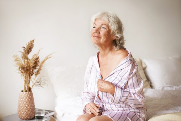 sensual gorgeous mature female with gray hair enjoying beautiful sunny morning in bedroom, sitting on bed, undressing. retired sexy caucasian woman in pajamas relaxing at home