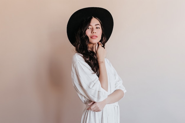 Sensual korean woman in black hat looking at camera. Studio shot of ecstatic asian model isolated on beige background.