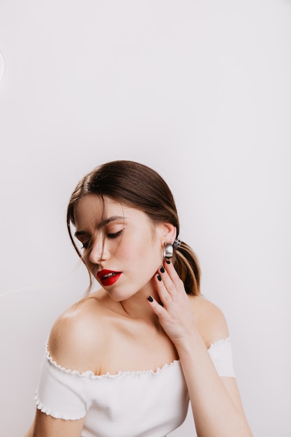 Sensual lady with plump red lips gently touches her neck, looking down. Shot of brown-haired girl in white top.