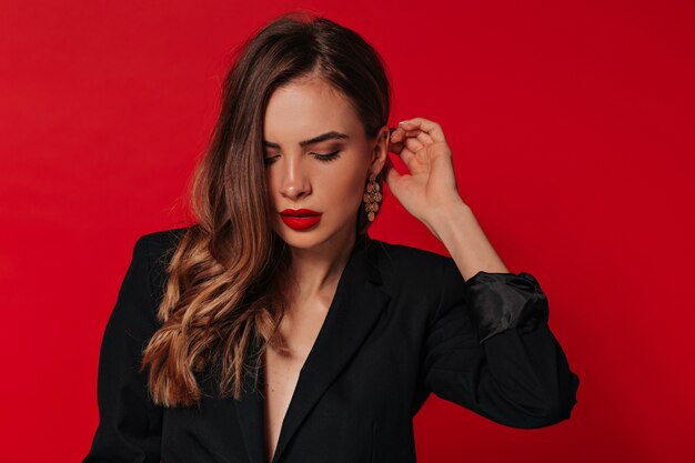 Sensual pretty woman with red lips wearing gold earrings and black jacket posing over red wall
