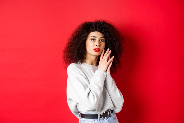 Sensual and romantic girl with curly hair and red lips, gently touching face with make-up, standing against red background.