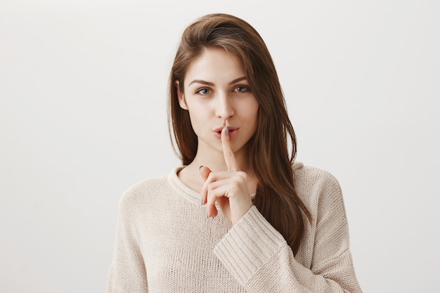 Sensual smiling woman shushing with finger pressed to lips, hiding secret