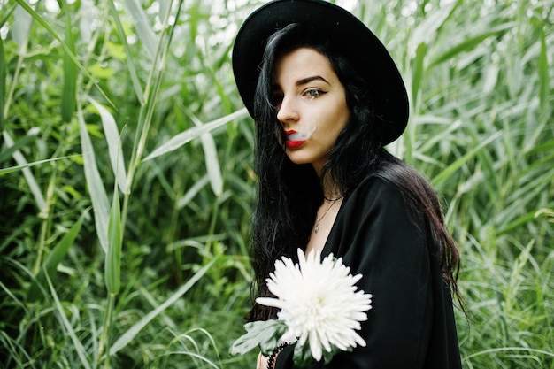 Sensual smoker girl all in black red lips and hat Goth dramatic woman hold white chrysanthemum flower and smoking on common reed