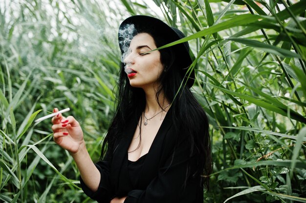 Sensual smoker girl all in black red lips and hat Goth dramatic woman smoking on common reed