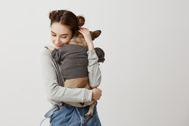 Sensual woman with childish hairstyle being mom of puppy while it lying on her shoulder with back. Feeling of care and love expressing by female pet owner.