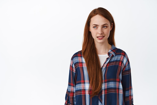 Sensual young redhead woman biting lip looking aside at logo with thoughtful coquettish face expression standing against white background