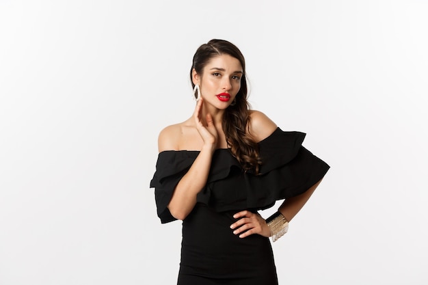 Sensual young woman in black dress, showing her earrings and looking sexy at camera, standing over white background