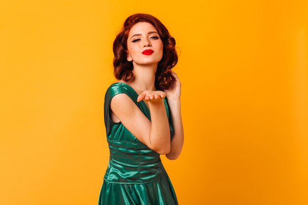 Sensual young woman in green dress sending air kiss. Front view of attractive girl with ginger wavy hair isolated on yellow space.