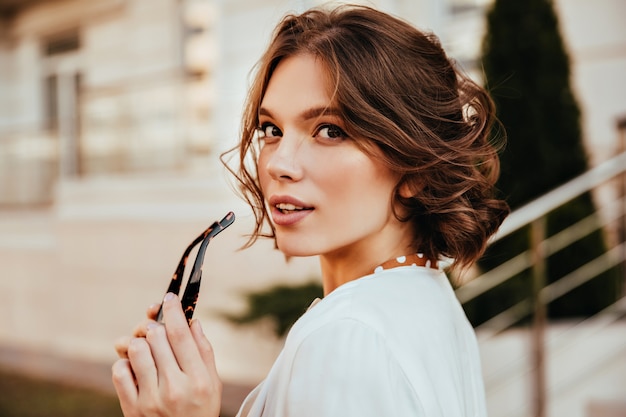 Sensual young woman in white blouse looking over shoulder. Positive elegant girl with short hair posing on the street.