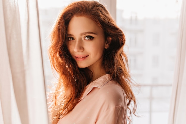 Free photo sensual young woman with ginger hair standing near window. indoor portrait of appealing white girl.