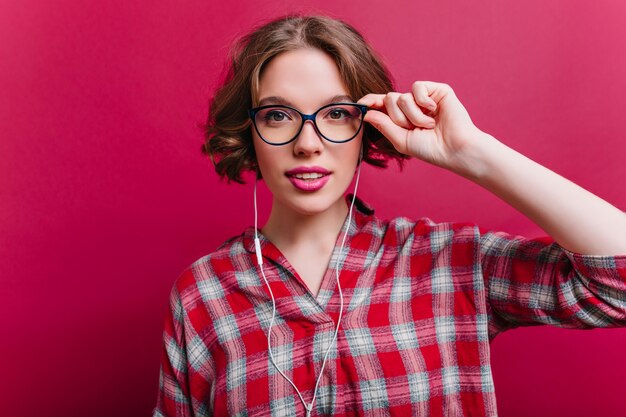 Sensual young woman with pink lips looking with interest and touching glasses.  charming curly girl posing in white earphones.