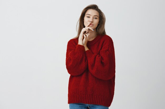 Sensuality, feminine and beauty concept. Portrait of charming flirty and sassy young sexy woman in red loose winter sweater holding index finger on lips hiding cheeky secret, shushing over white wall